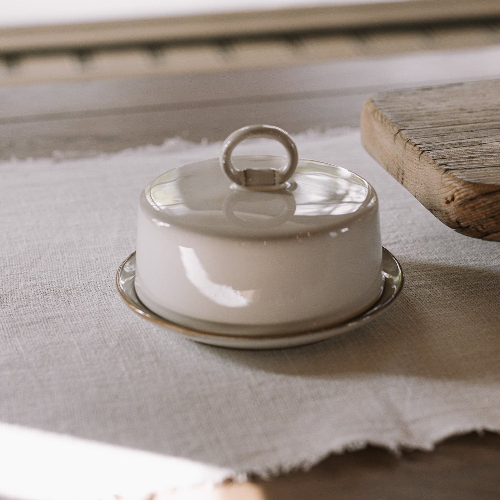 Off white butter dish with lid and loop to open on neutral wooden table with white table cloth.