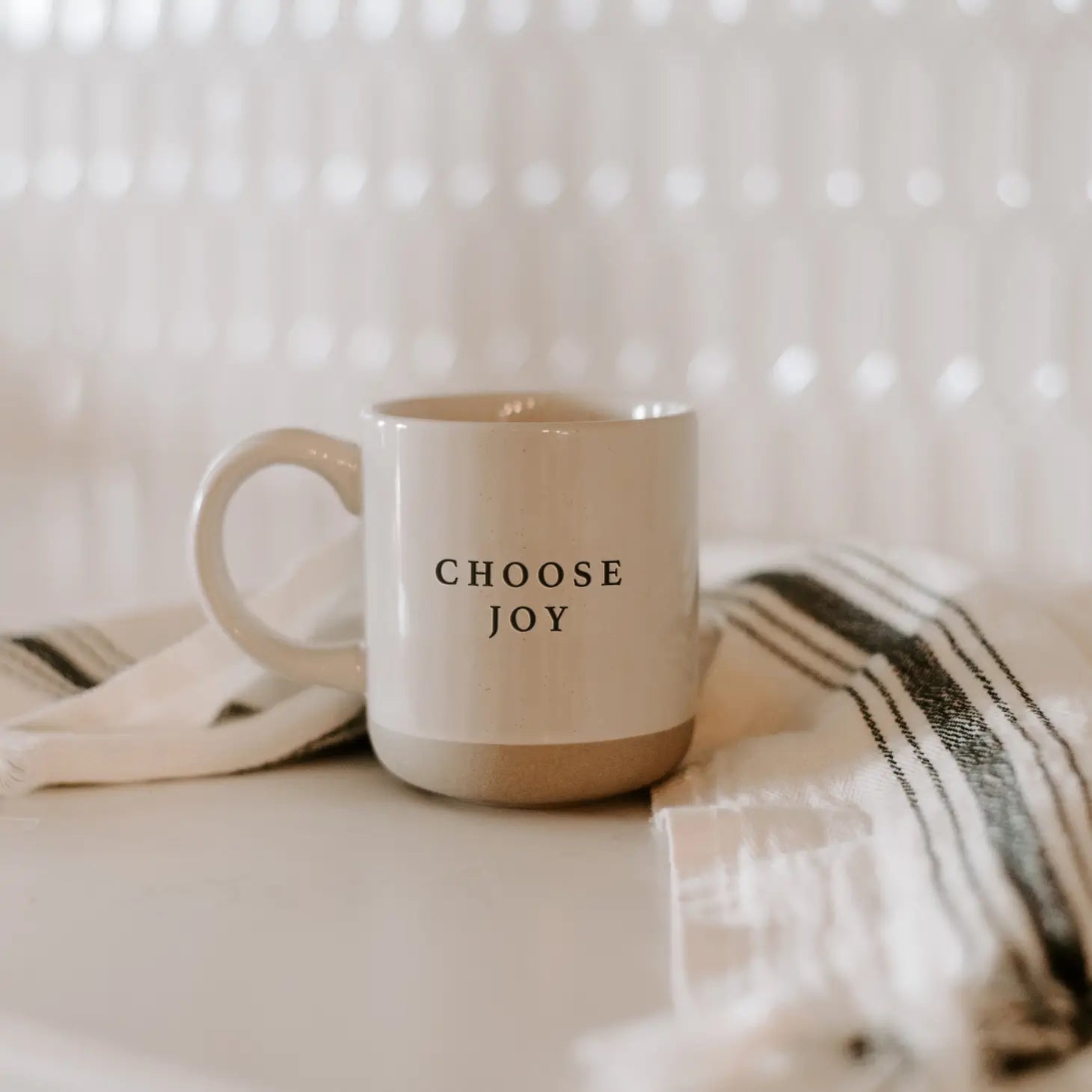 White and brown stoneware mug with 'choose joy' in black lettering, on white and black tea towel.