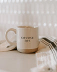 White and brown stoneware mug with 'choose joy' in black lettering, on white and black tea towel.