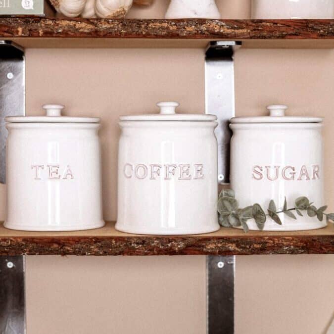 Ceramic white tea coffee sugar canisters on wooden shelf with green stem.