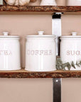 Ceramic white tea coffee sugar canisters on wooden shelf with green stem.