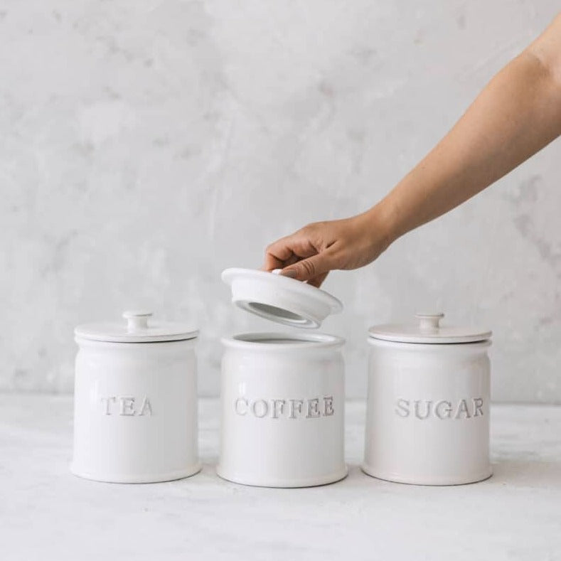 White ceramic tea coffee sugar canisters on grey background with one being opened.