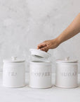 White ceramic tea coffee sugar canisters on grey background with one being opened.