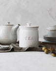 White ceramic tea storage jar with tea pot and crockery behind on tablecloth.
