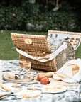 Round wicker picnic basket open on picnic blanket with food and wine.