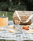 Round wicker picnic basket closed on checked picnic blanket with outside fire and food and drinks.