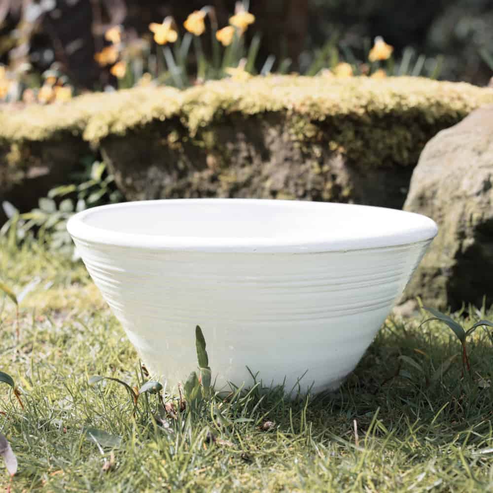 White ridged ceramic bowl outside on the grass with stones and flowers in background.