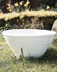 White ridged ceramic bowl outside on the grass with stones and flowers in background.