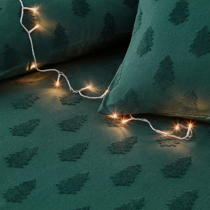 Close up of green Christmas bedding with trees, with duvet and cushions with fairy lights on top.