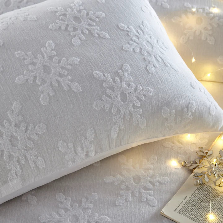 Close up of cushion on white Christmas bedding set with snowflakes, with fairy lights and book.