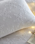 Close up of cushion on white Christmas bedding set with snowflakes, with fairy lights and book.