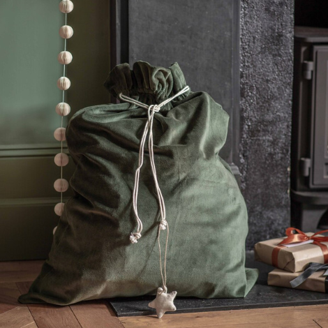 Forest green velvet Christmas sack with drawstring and star decoration, filled with presents in front of a fireplace.