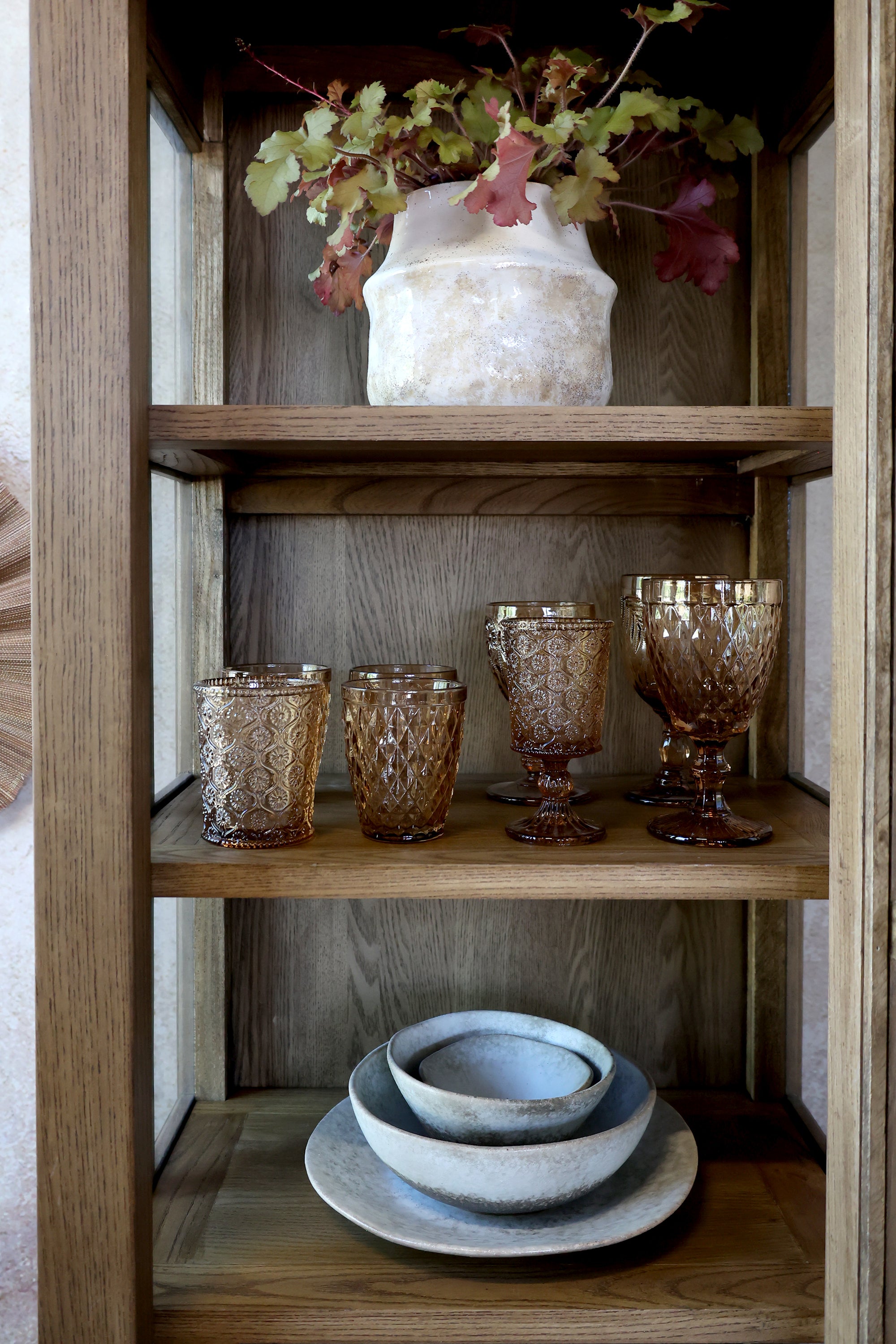 Close up of shelving on natural wood display cabinet, with amber glasses, a white vase and dinnerware.