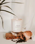 Fresh coffee soy candle in matte white jar, on wooden plank with spilled beans out of terracotta jar.