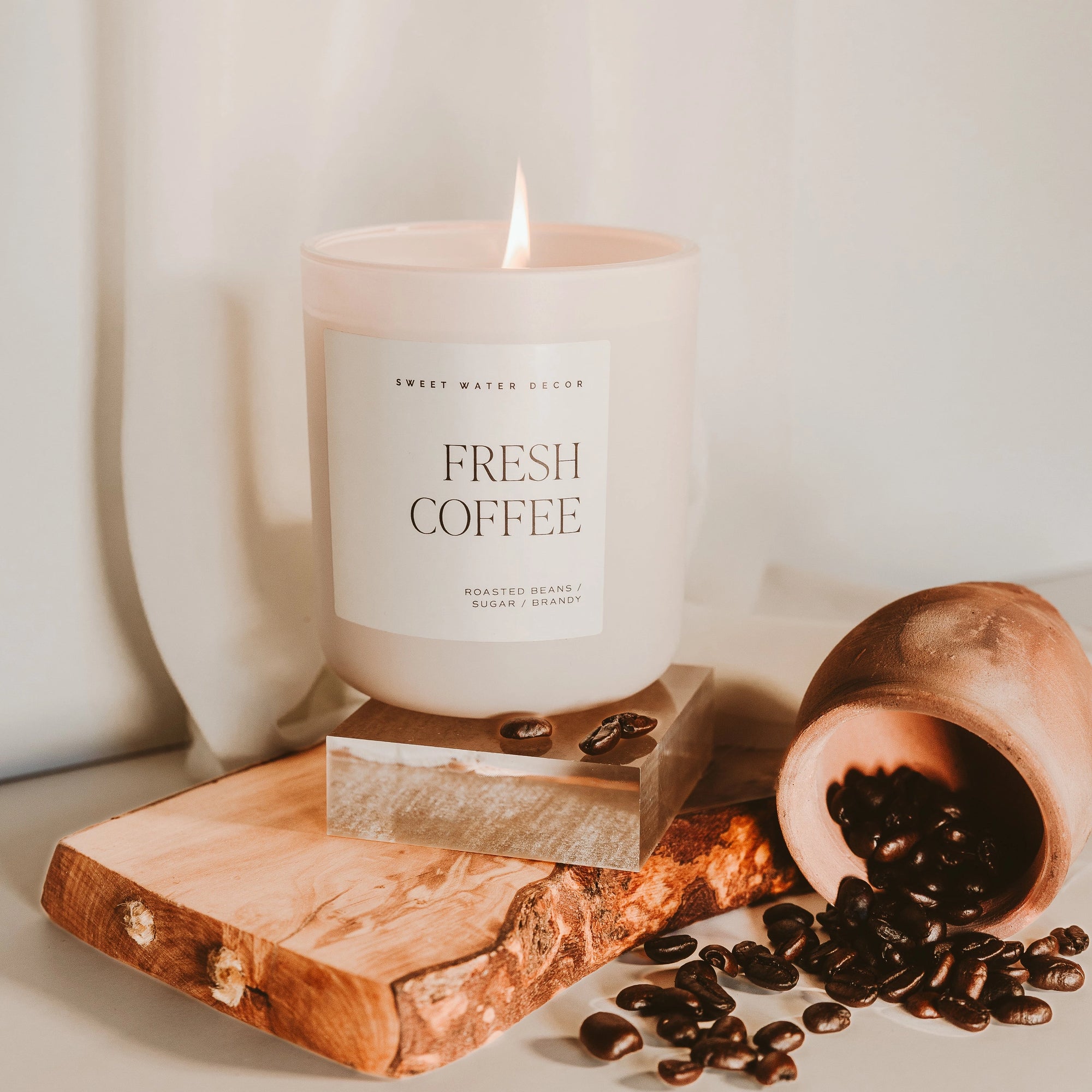 Fresh coffee soy candle in matte white jar, lit on a wood plaque with coffee beans spilled over.
