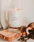 Fresh coffee soy candle in matte white jar, lit on a wood plaque with coffee beans spilled over.