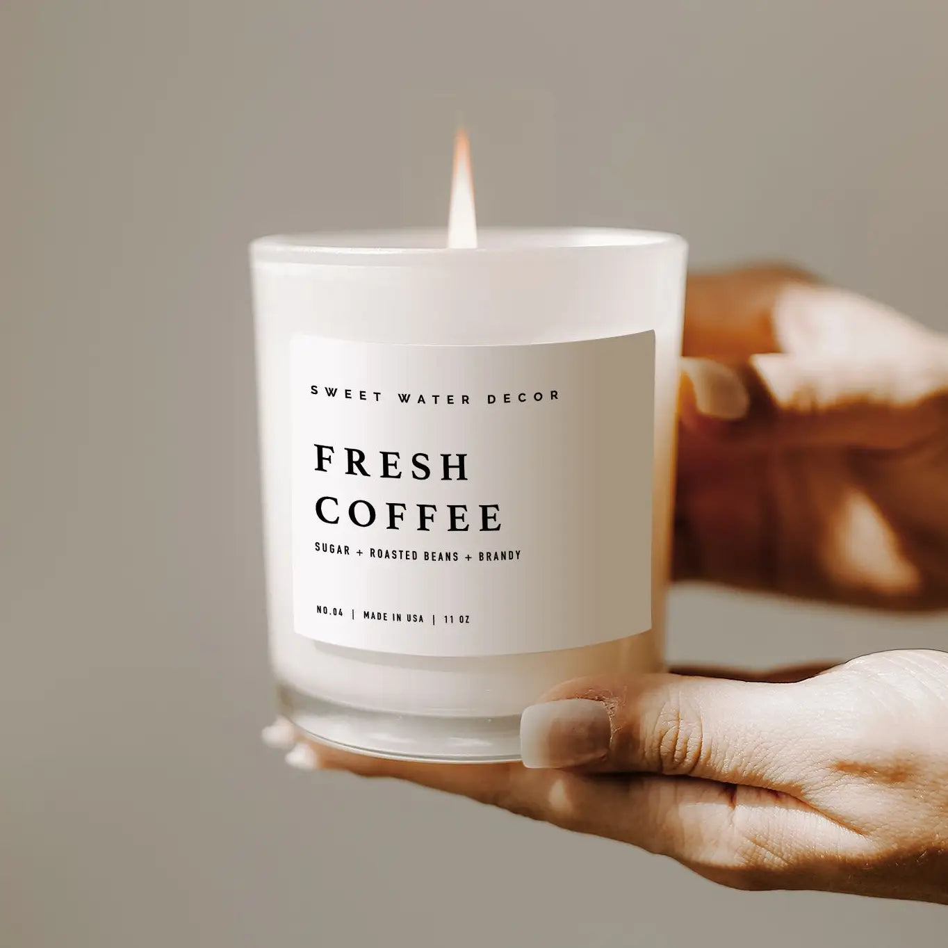 Fresh coffee soy candle in white jar, lit and held up in two hands.