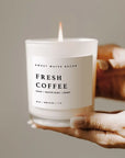 Fresh coffee soy candle in white jar, lit and held up in two hands.