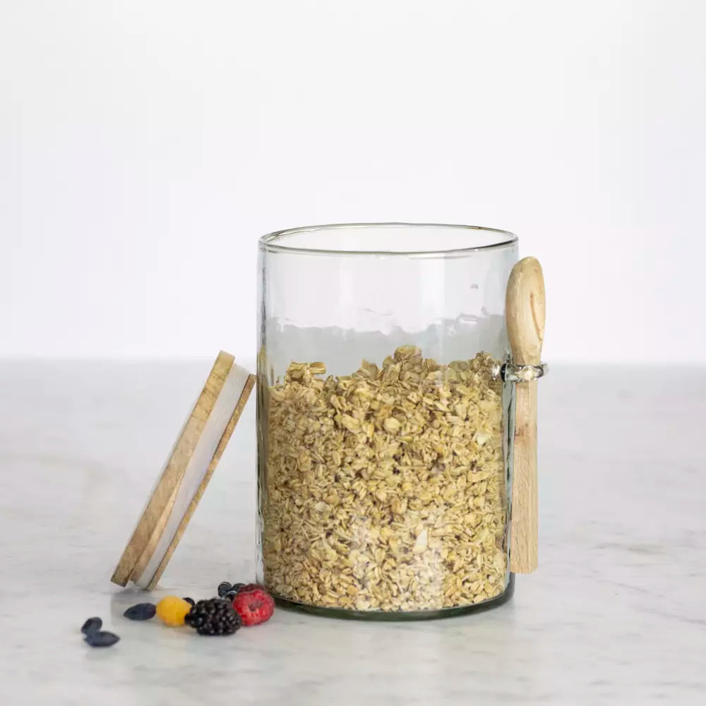 Hammered glass storage jar with wooden lid and spoon, filled with oats with berries on the side.