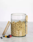 Hammered glass storage jar with wooden lid and spoon, filled with oats with berries on the side.