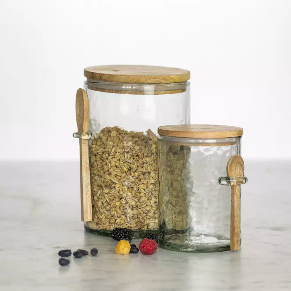 Two hammered glass storage jars with wooden lids and spoons, one filled with oats and berries.