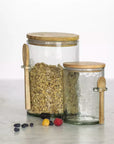 Two hammered glass storage jars with wooden lids and spoons, one filled with oats and berries.