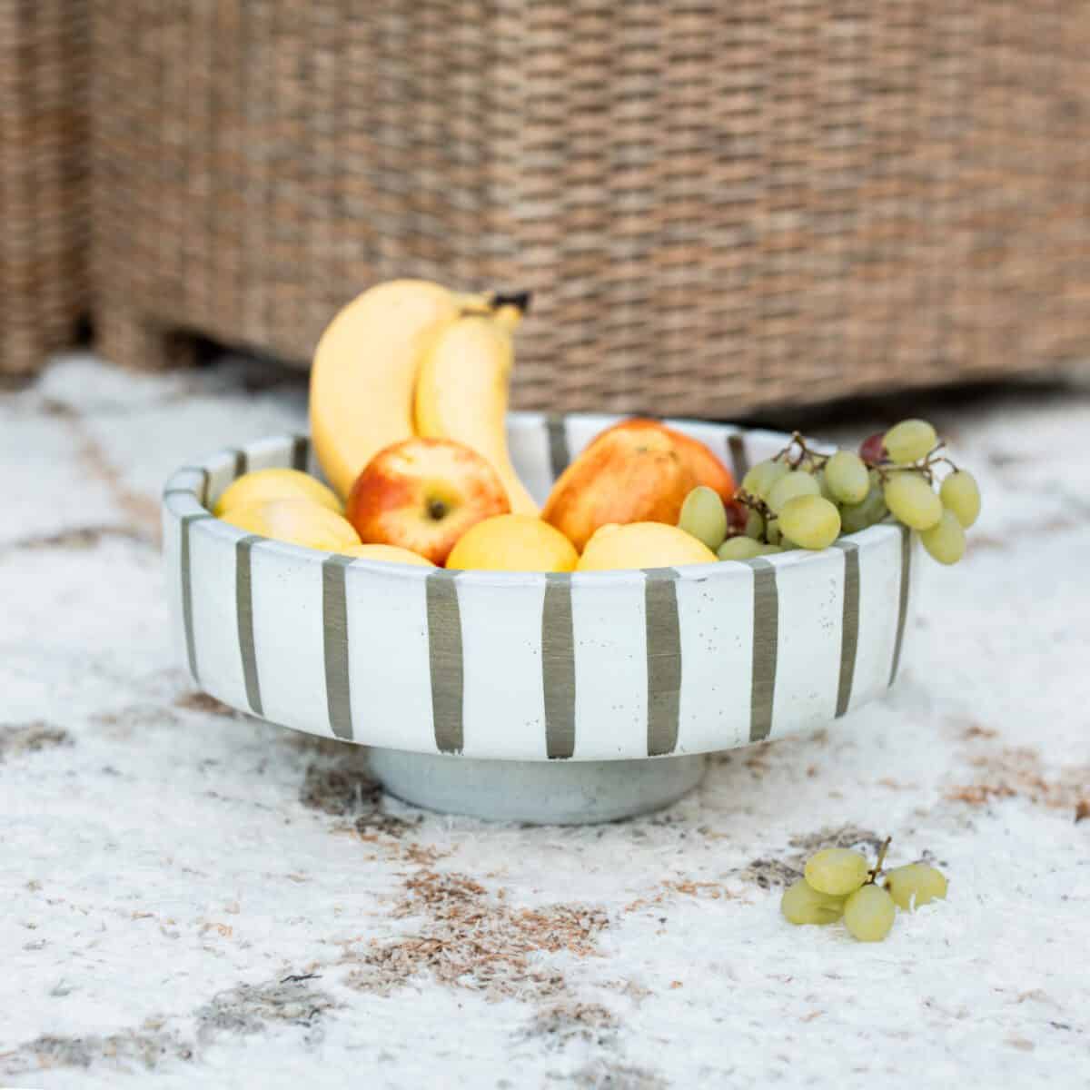 Striped ceramic fruit bowl filled with fruit outside on floor.