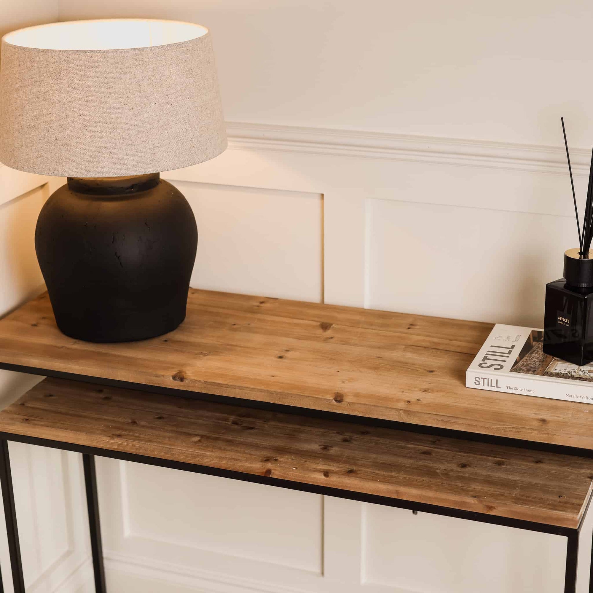 Two wooden nest console tables with switched on lamp, white book and black diffuser from above view.
