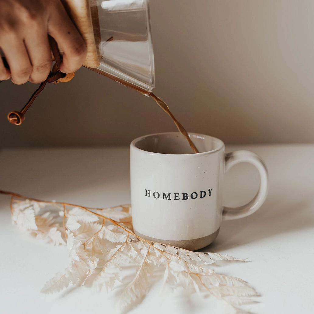 White and brown stoneware mug with black lettering 'homebody', on a white table with coffee being poured in.
