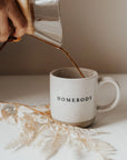 White and brown stoneware mug with black lettering 'homebody', on a white table with coffee being poured in.