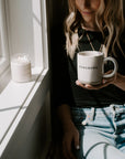 White and brown stoneware mug with black lettering 'homebody', held underneath next to a window.