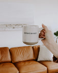 White and brown stoneware mug with black lettering 'homebody', held up by the handle in front of orange sofa.