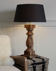 Tall column wooden lamp on side table with black shade, lit.