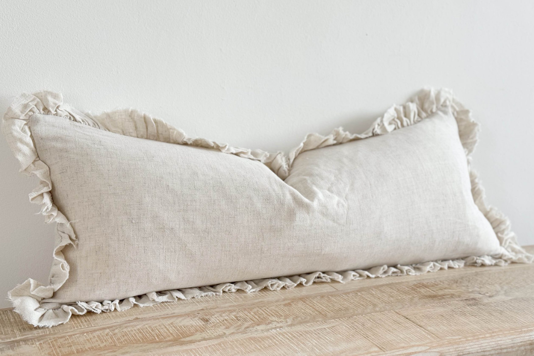 Sand coloured bolster cushion with ruffled edge on wooden surface.