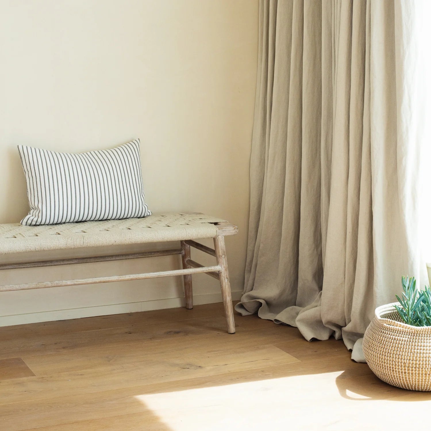 A neutral room with sunlight coming through the window. The wooden bench is against the wall with a striped cushion.