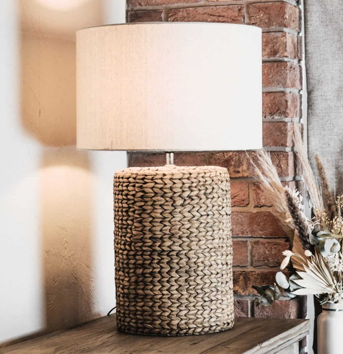 Rope effect tall lamp with light linen shade on wooden console, in front of brick wall.