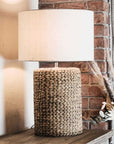 Rope effect tall lamp with light linen shade on wooden console, in front of brick wall.