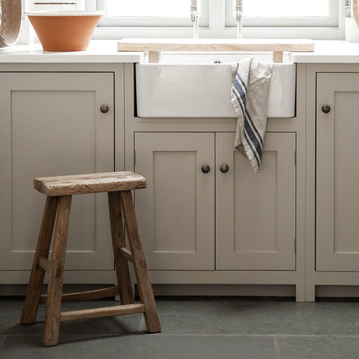 A neutral shaker style kitchen with white sink and small rustic wooden stool.