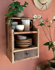 Small wooden shelf with drawer hung on wall with crockery on and plants surrounding.