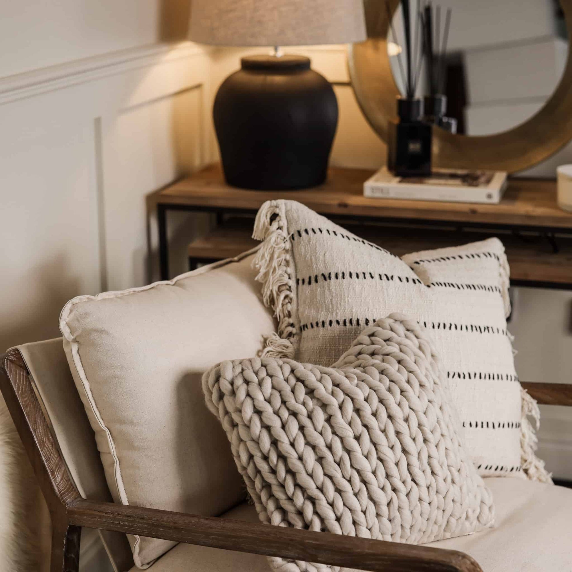 Two neutral cushions on an armchair with wooden console in background and black lamp.