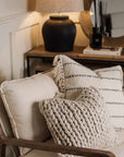 Two neutral cushions on an armchair with wooden console in background and black lamp.