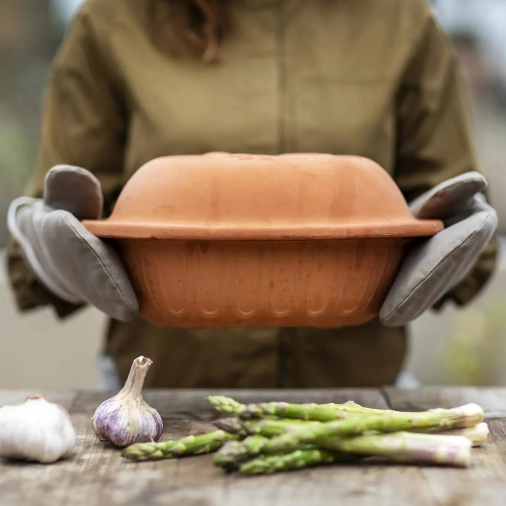 Organic clay oven gloves holding terracotta skillet with veg underneath.