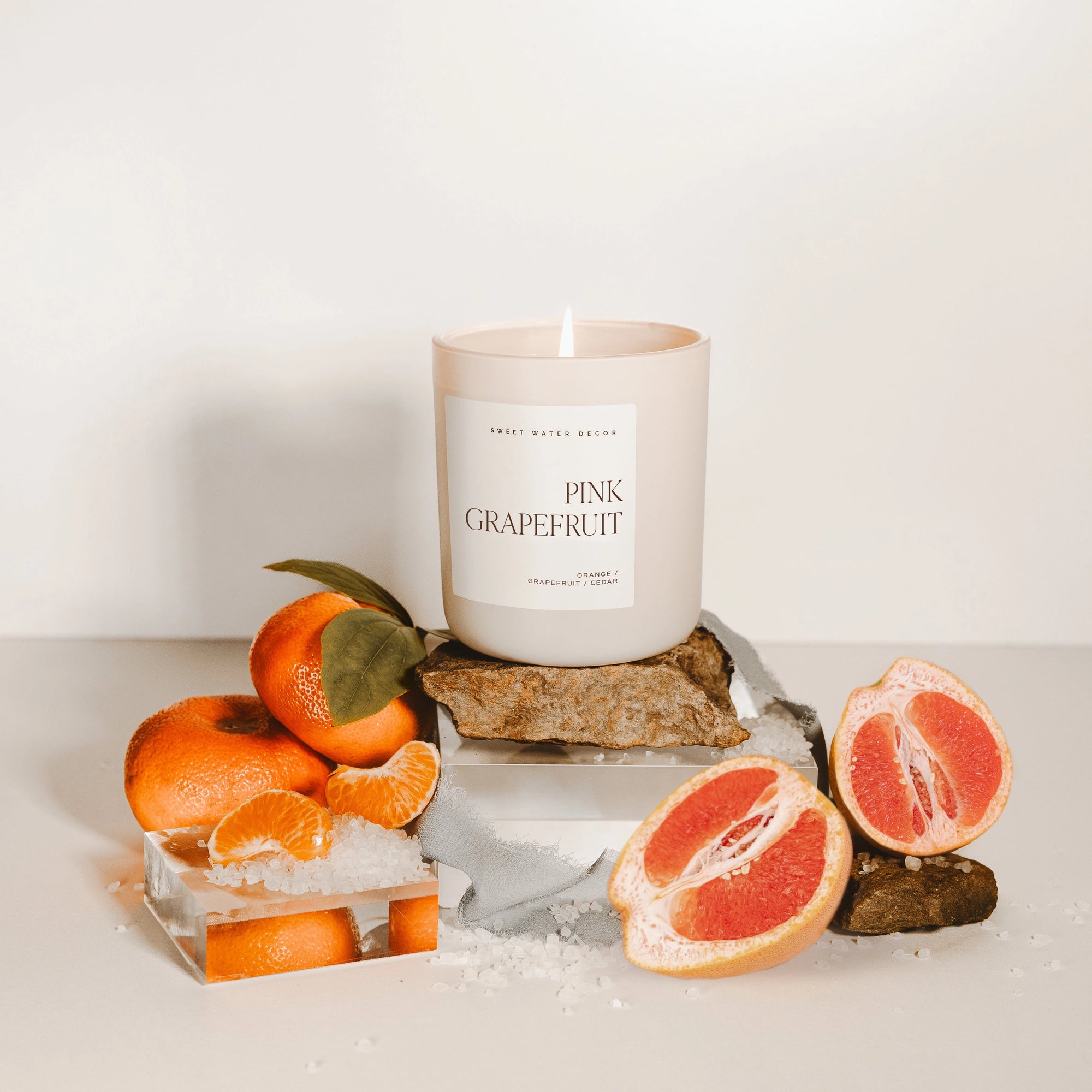 Pink grapefruit soy candle on a wood plaque, lit, surrounded by fruits and salt.