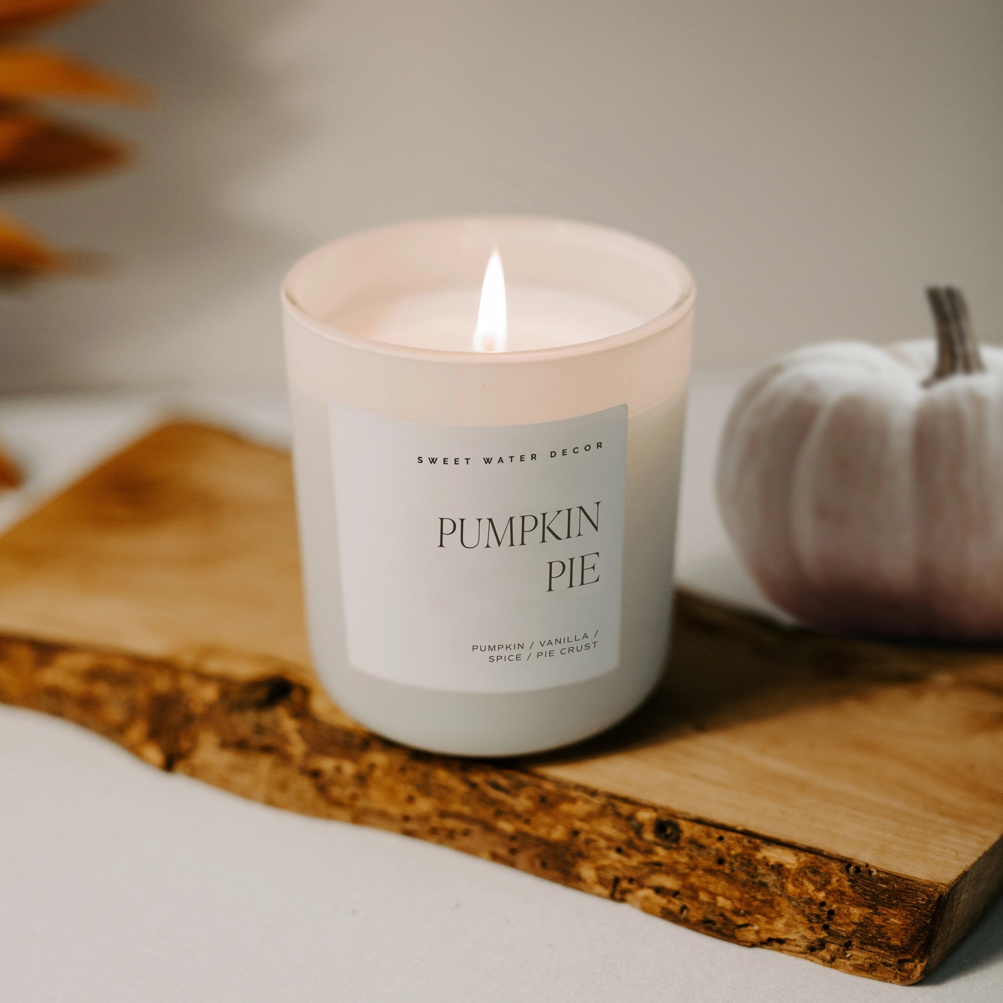 White soy candle lit on a wooden plank with white pumpkin decoration behind.