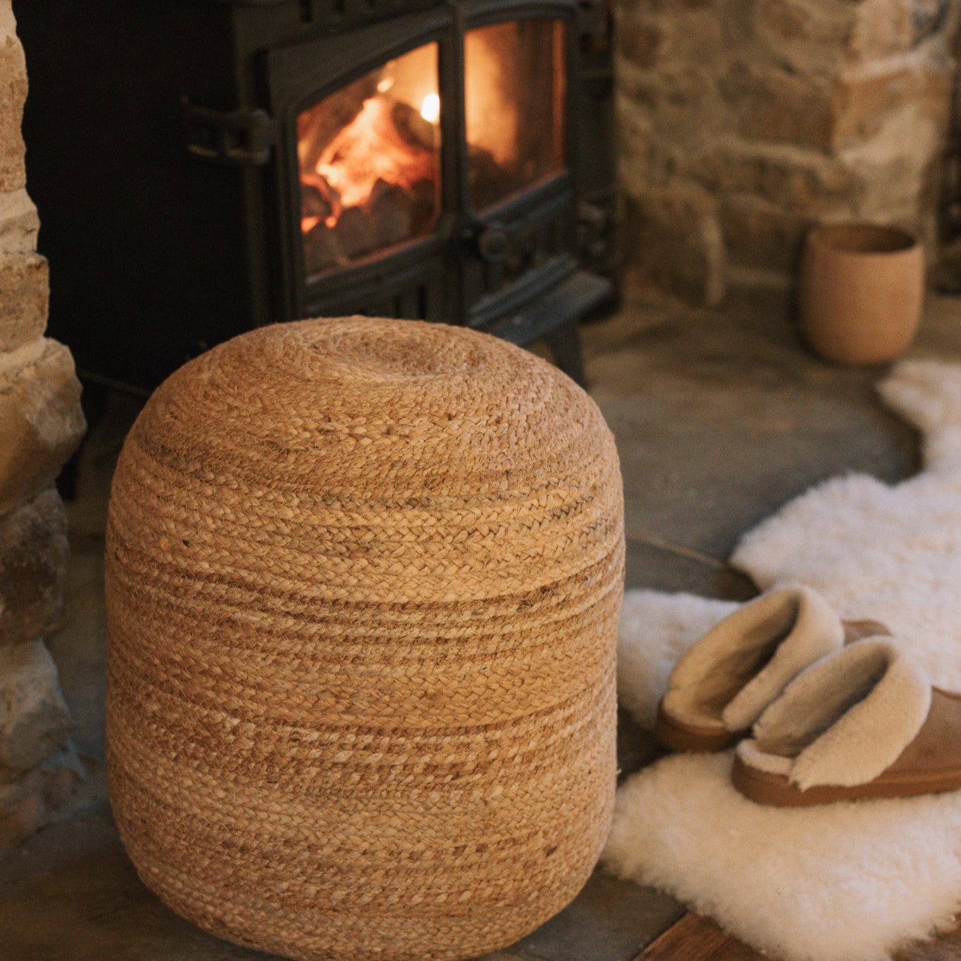 Round jute pouffe in front of wood burning stove with slippers and sheepskin rug.