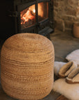 Round jute pouffe in front of wood burning stove with slippers and sheepskin rug.