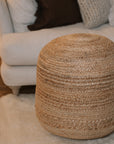 Round jute pouf in front of sofa on white rug.