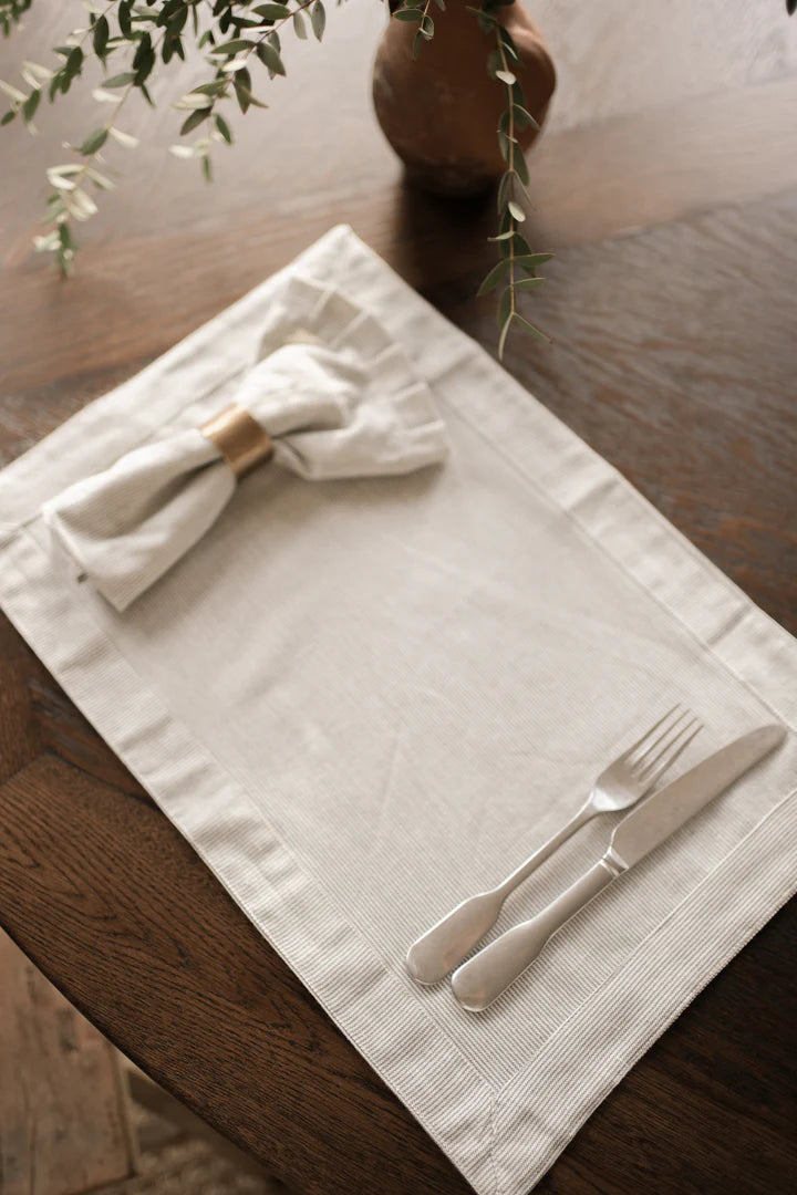 Pinstripe napkin in a brass napkin ring, on matching placemat with cutlery on dark wood dining table.