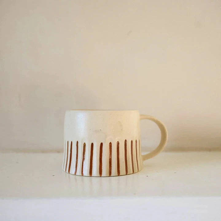 Off white handmade mug, with rust coloured hand painted stripes, on a white shelf.