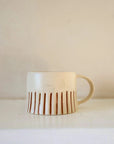 Off white handmade mug, with rust coloured hand painted stripes, on a white shelf.